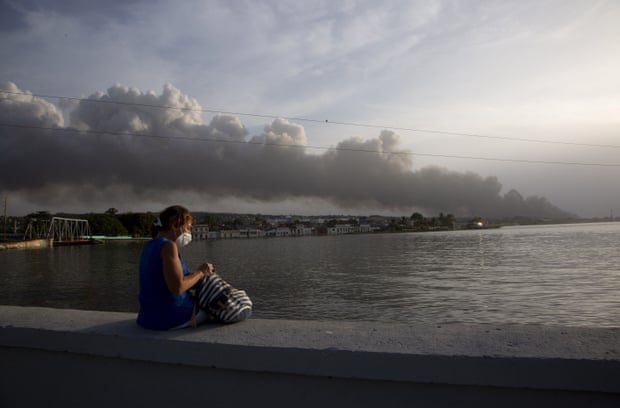 A resident sits on the Malecon sea wall as smoke rises in the background from the fire.
